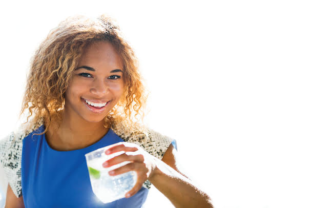 Attractive young African American woman sitting by a bright sunny window, drinking a glass of water.